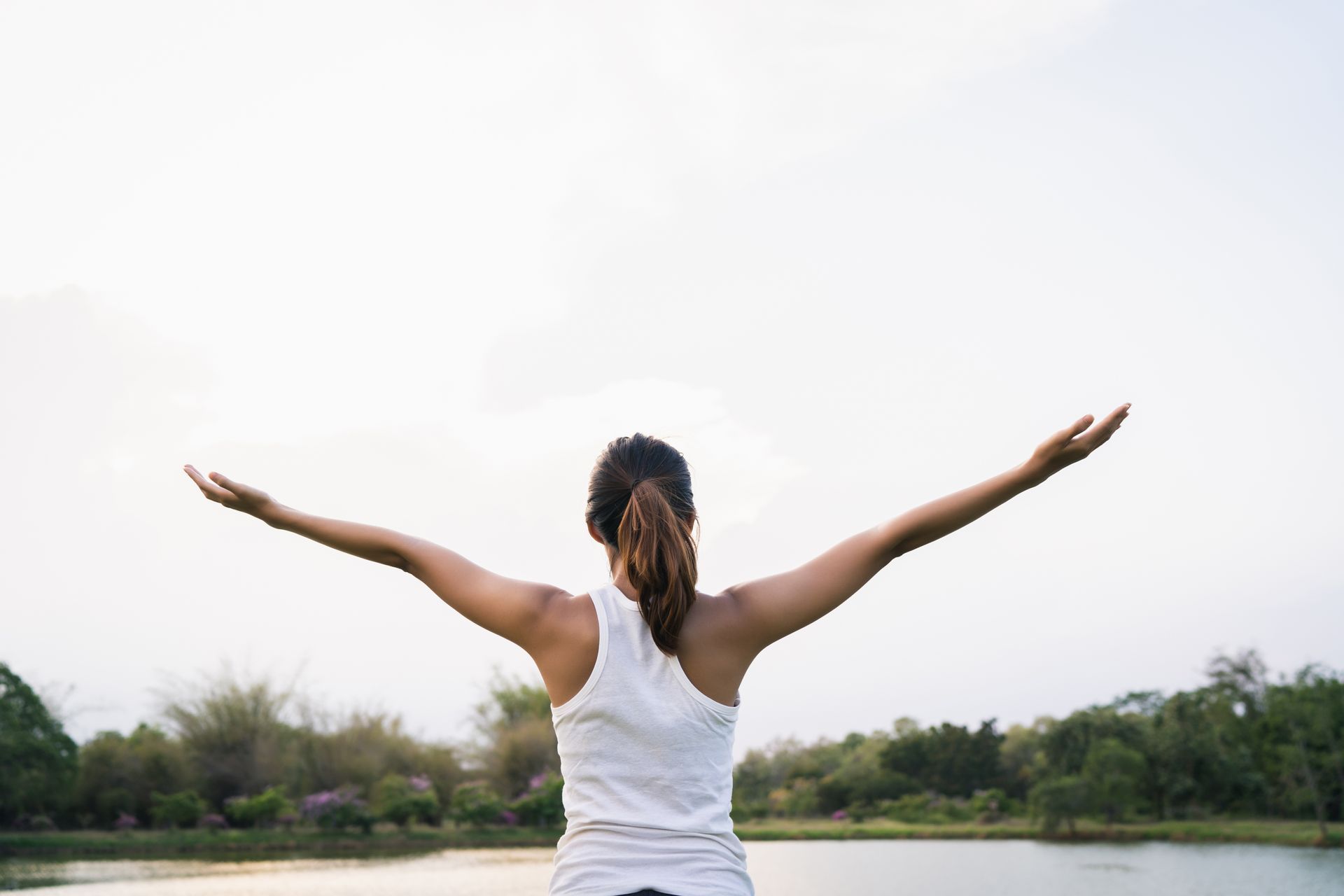 A woman is standing in front of a lake with her arms outstretched.