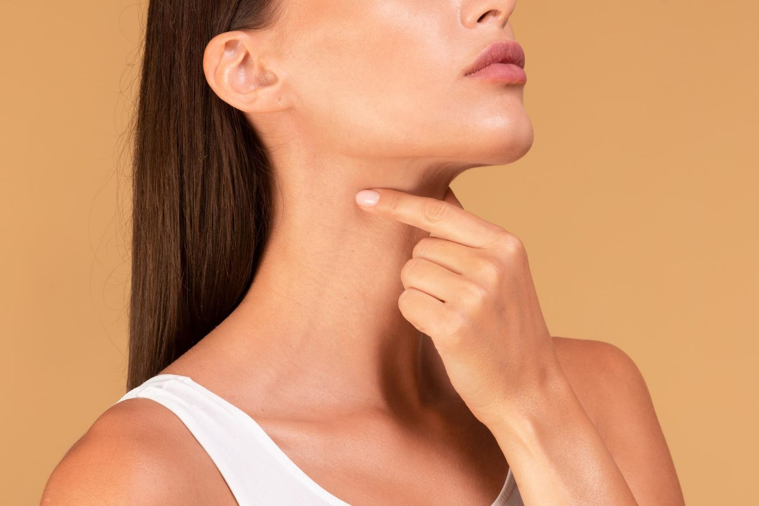 Woman touching her neck, possibly checking for a lump, wearing a white top. Beige background.