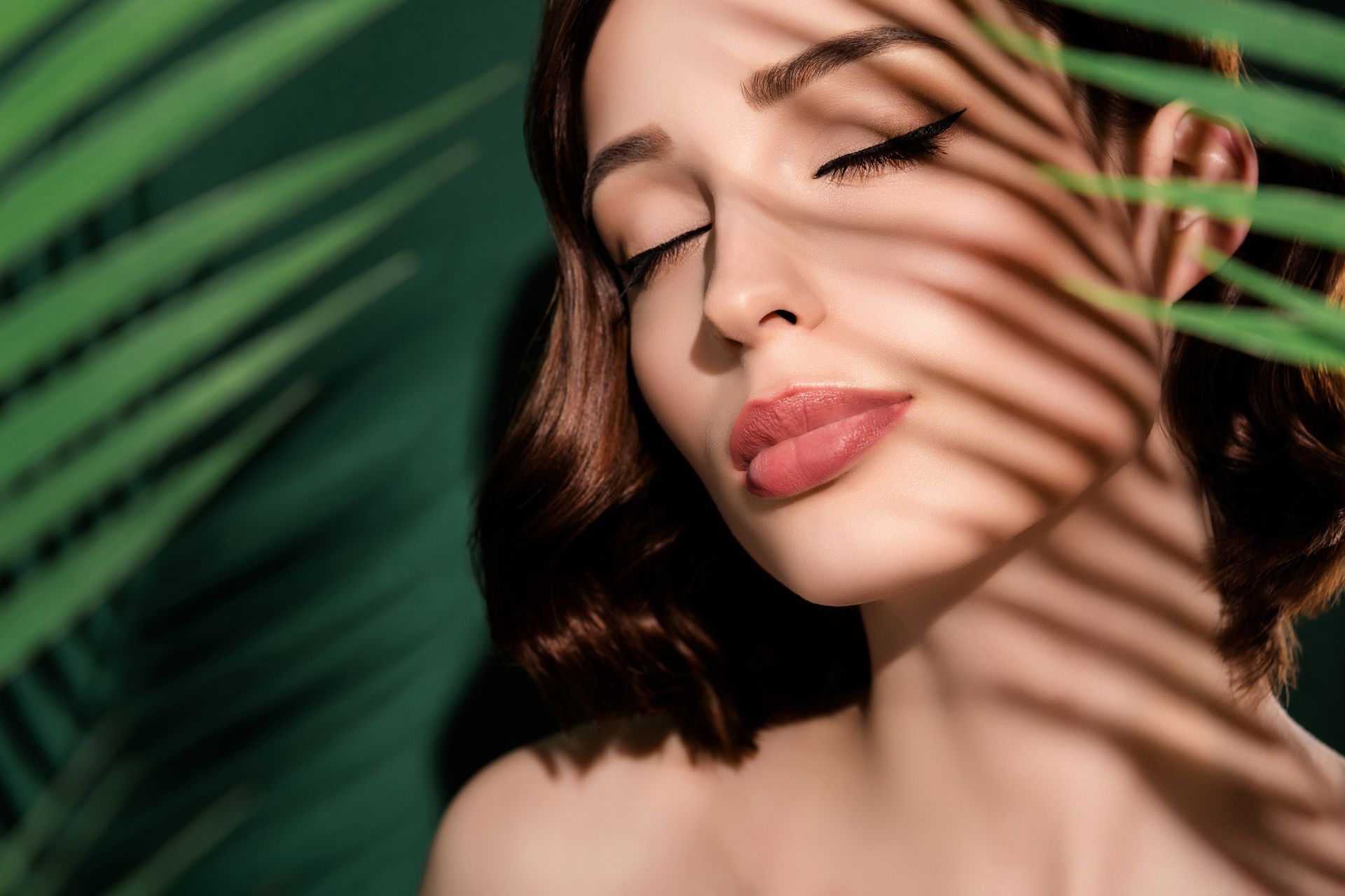 A woman with her eyes closed is standing in front of a palm tree leaf.