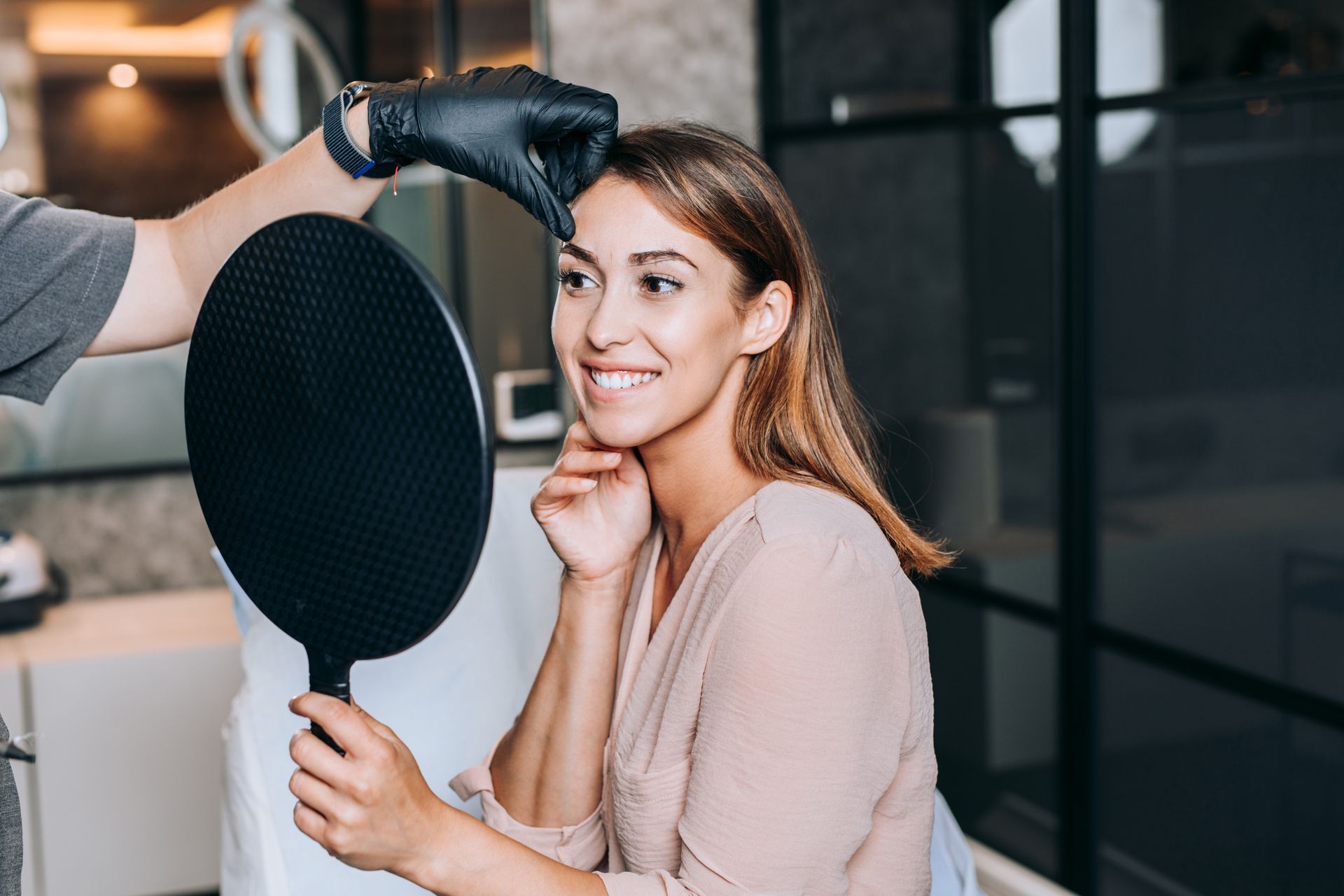 A person in black gloves examining the eyebrow of a smiling woman who holds a round black mirror in a salon.