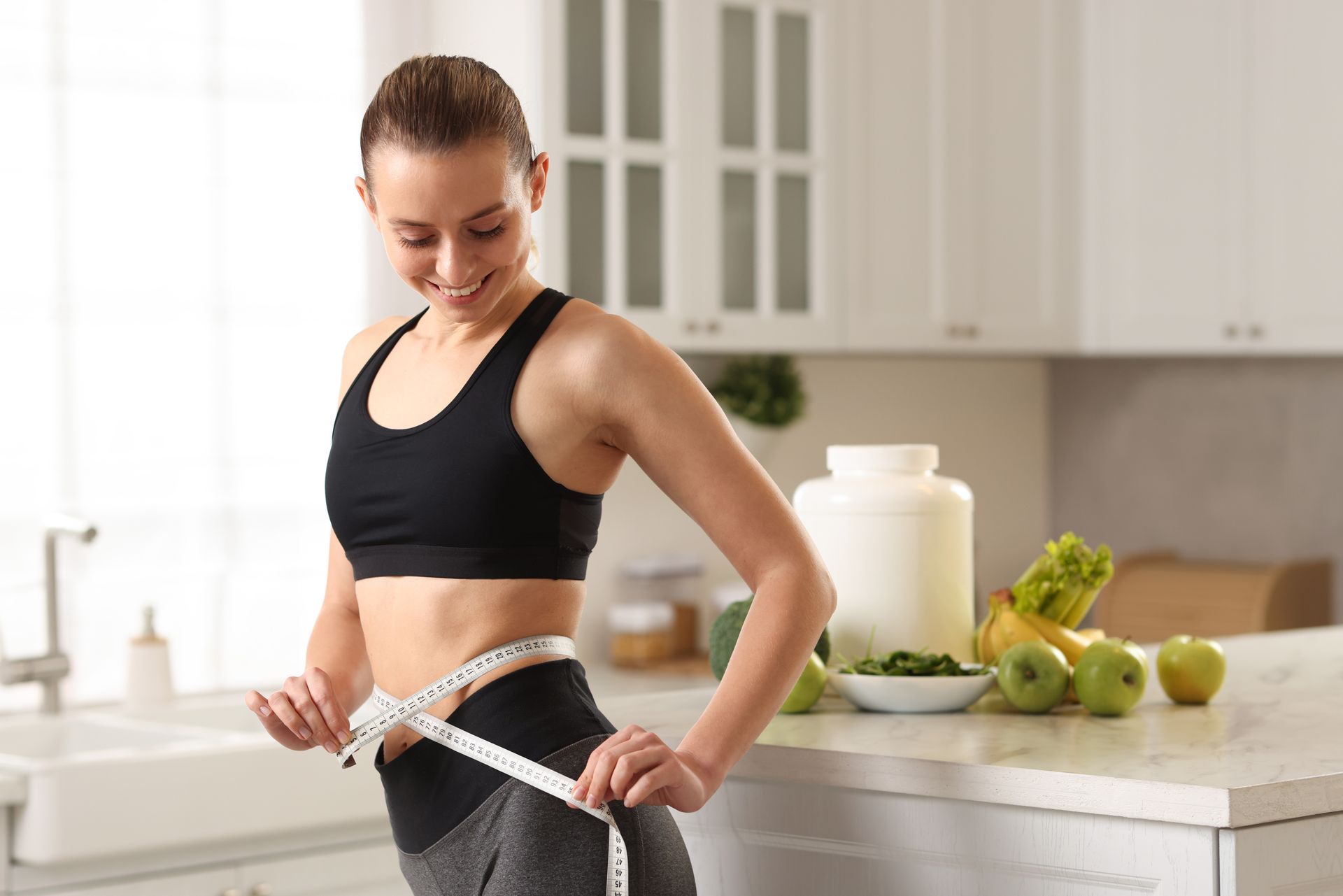 Woman in workout clothes measuring waist with a smile in a kitchen with fruit and protein powder.