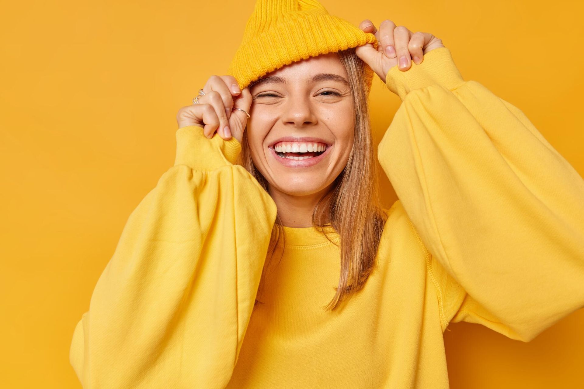 Woman in yellow outfit smiles, pulling on yellow beanie, against yellow backdrop.