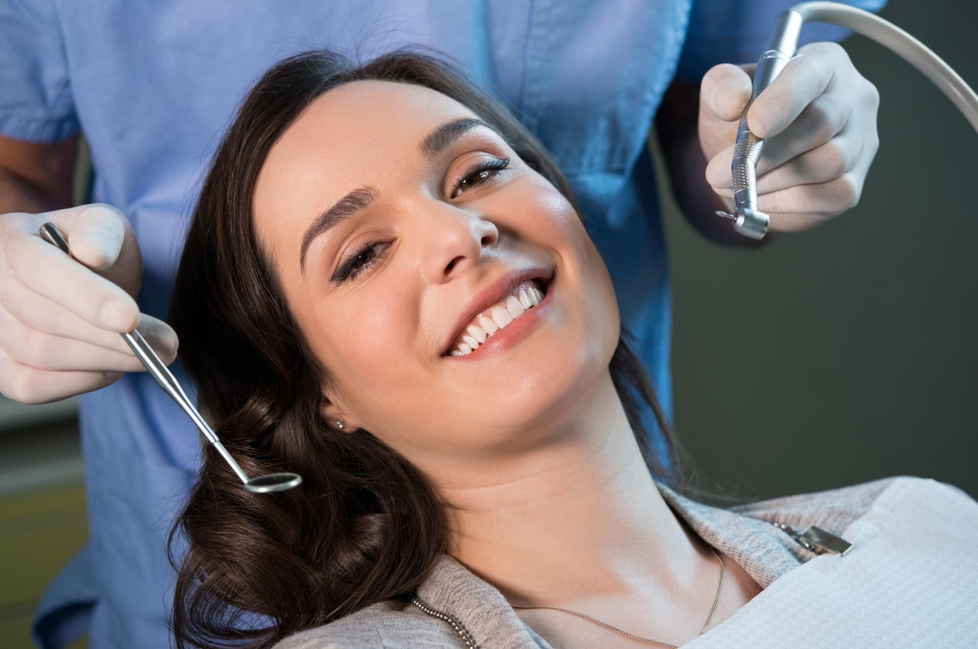 Woman in dentist's chair smiles at the camera, with dentist holding tools.