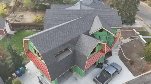 Aerial view of a house under construction with a dark gray roof and green insulation, a car parked in front.