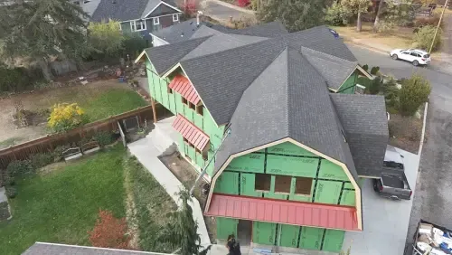 Barn-shaped house under construction with a dark gray roof and green siding. Red awnings cover windows.
