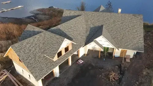 Aerial view of a house under construction with a gray roof and partially finished walls near a body of water.