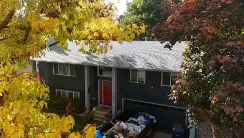 Gray house with red door, surrounded by trees with yellow and red leaves, dumpster in driveway.