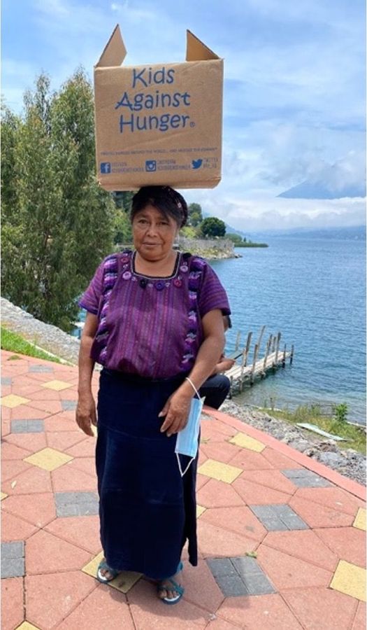 A poor Guatemalan woman with a cardboard box full of food on her head that says kids against hunger.