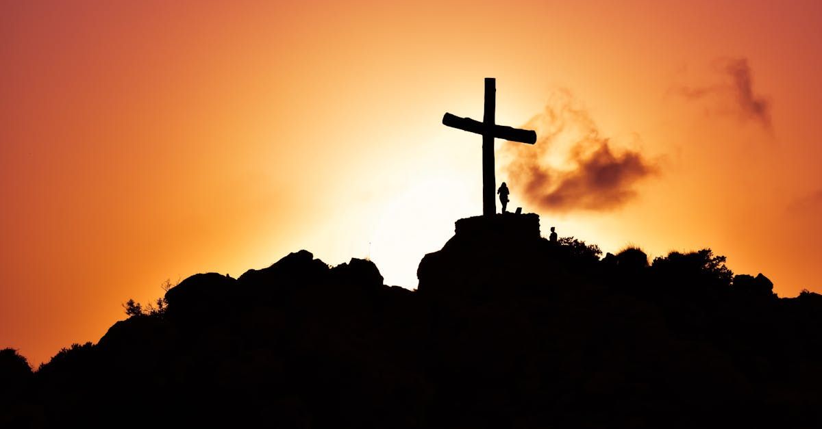 A silhouette of a man standing beside a cross on a rocky mountain top as the sun rises on the horizon.