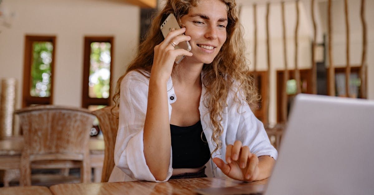 A woman is sitting at a table talking on a cell phone in front of a laptop computer.