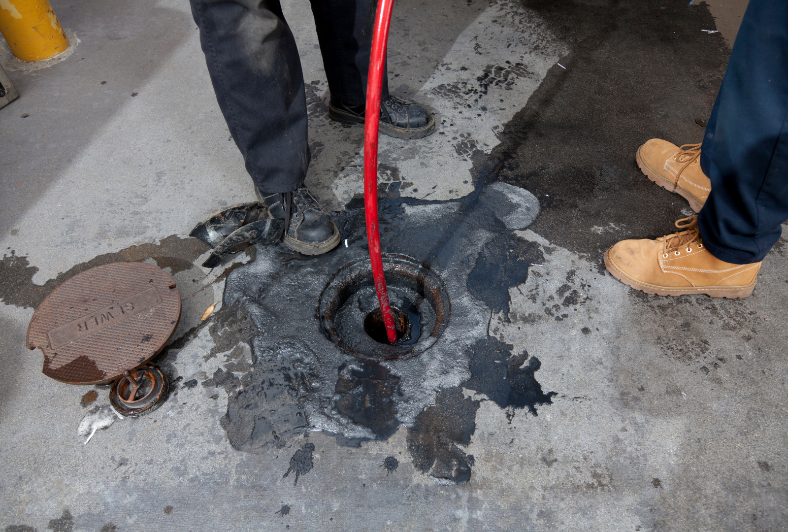 Workers clearing a clogged manhole with a red hose on a wet concrete street.