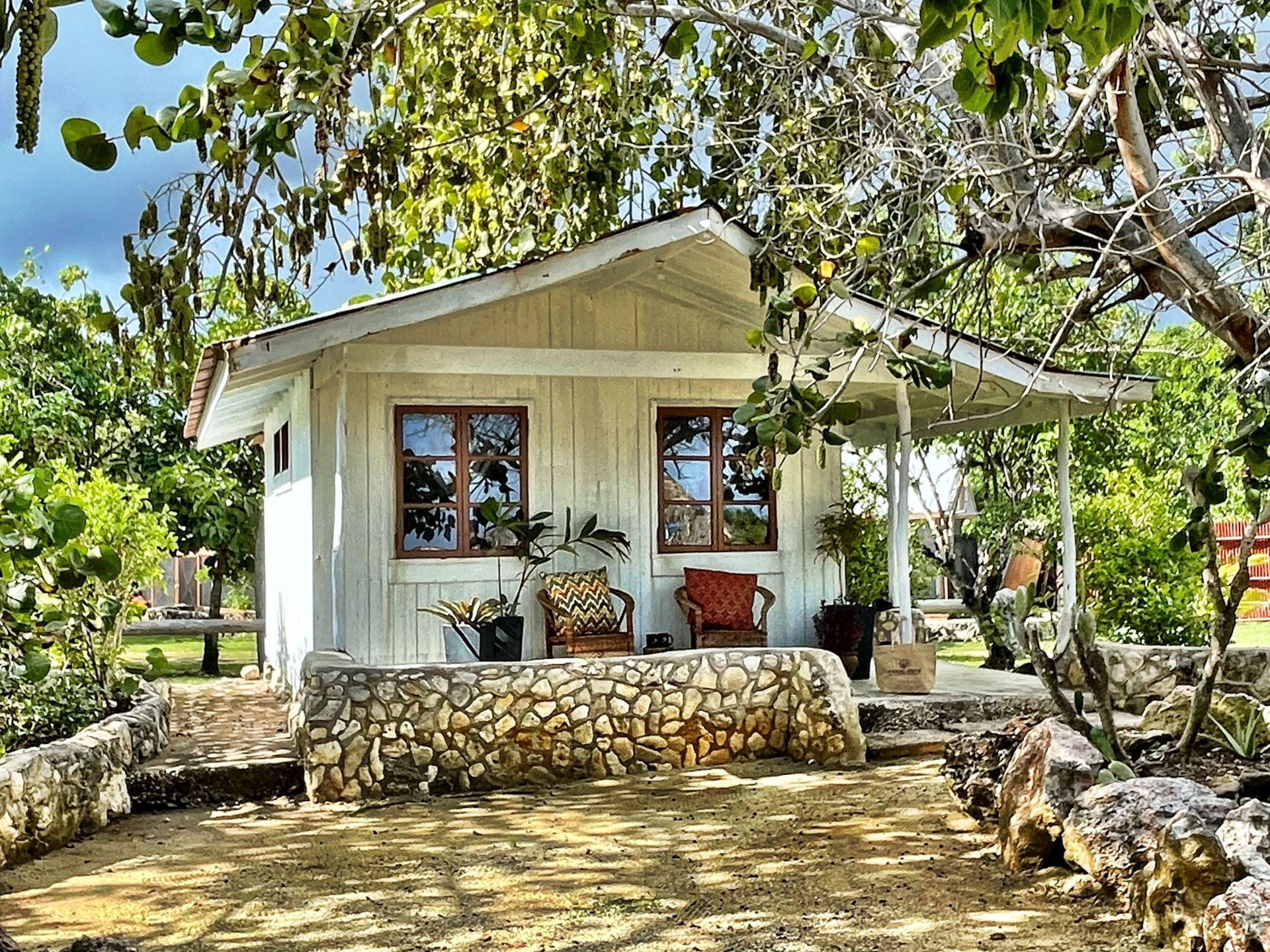 A small white house with a porch and chairs is surrounded by trees.