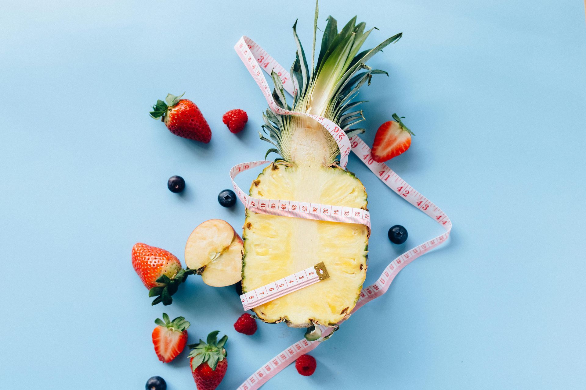 Pineapple and berries with measuring tape on a blue background, representing healthy eating and weight loss.