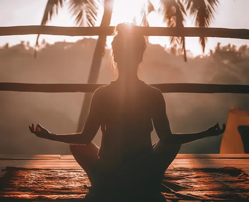 A woman is sitting in a lotus position while the sun shines through the palm trees.