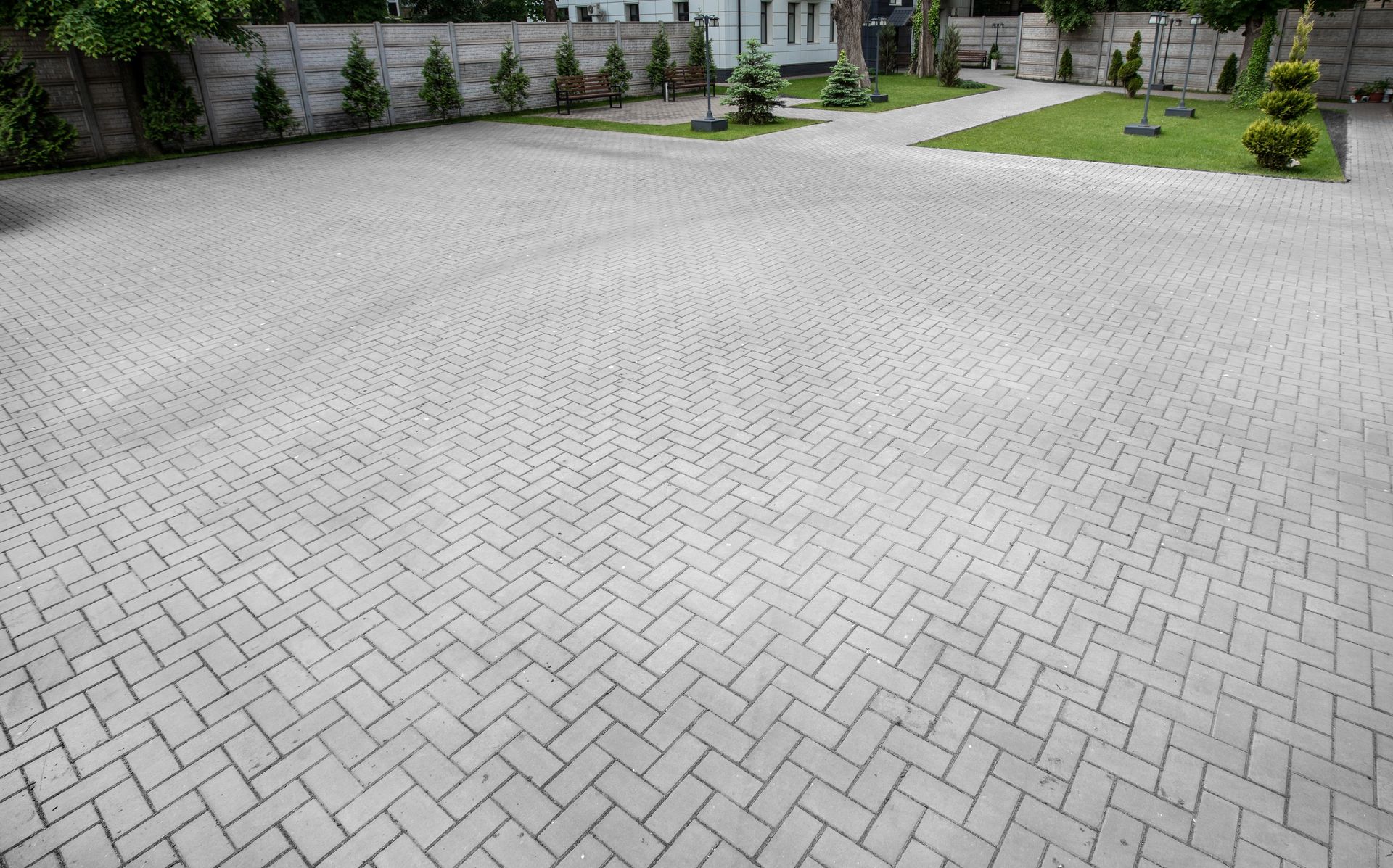 A wide, gray paved driveway with a herringbone brick pattern leading toward a grassy area with small shrubs and a wall.