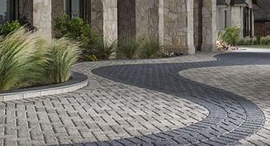 A curved driveway featuring light grey rectangular pavers bordered by a contrasting dark charcoal wavy path.