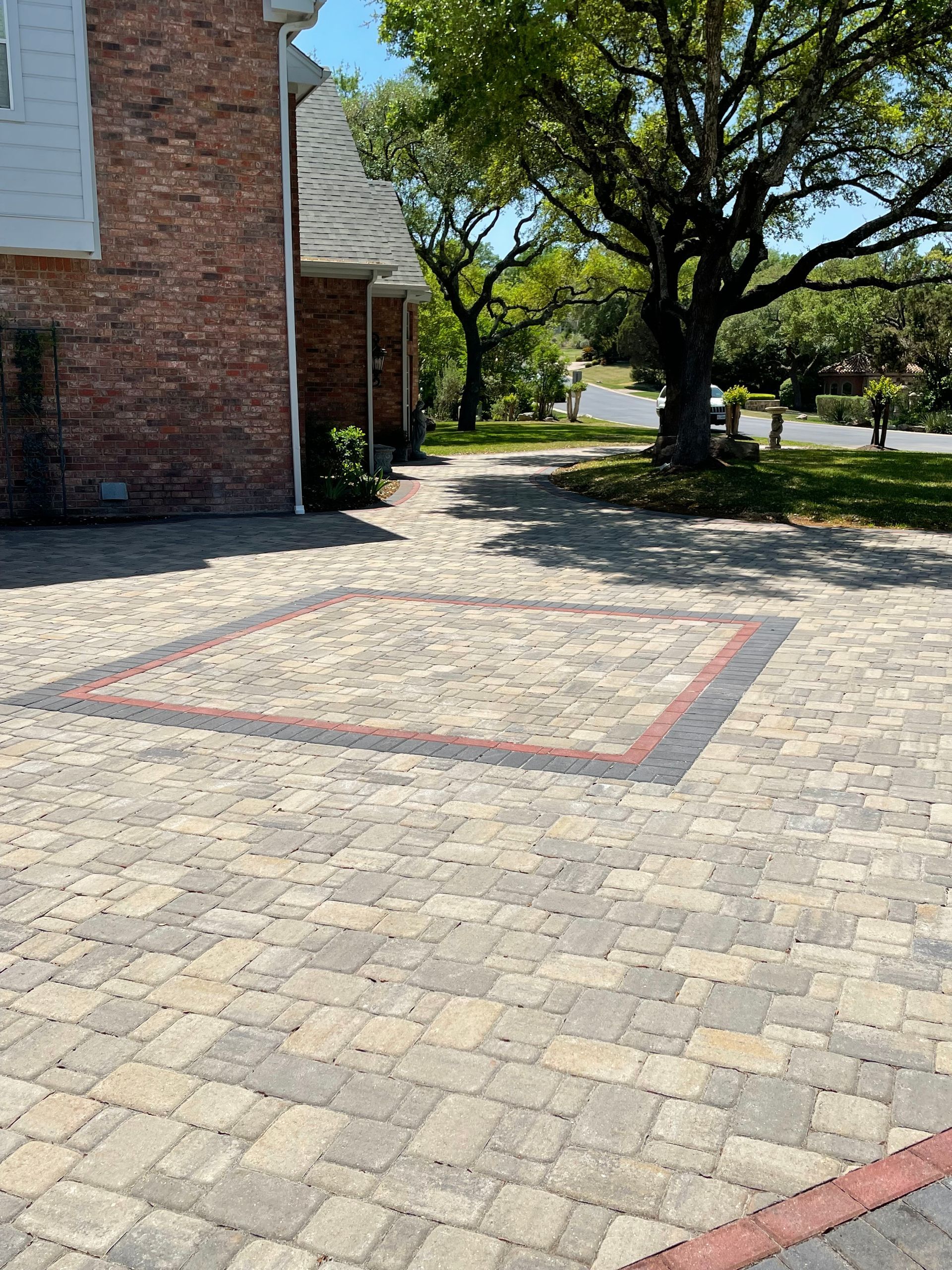 A paved driveway featuring a large square pattern with reddish-brown borders set against a brick house and green trees.
