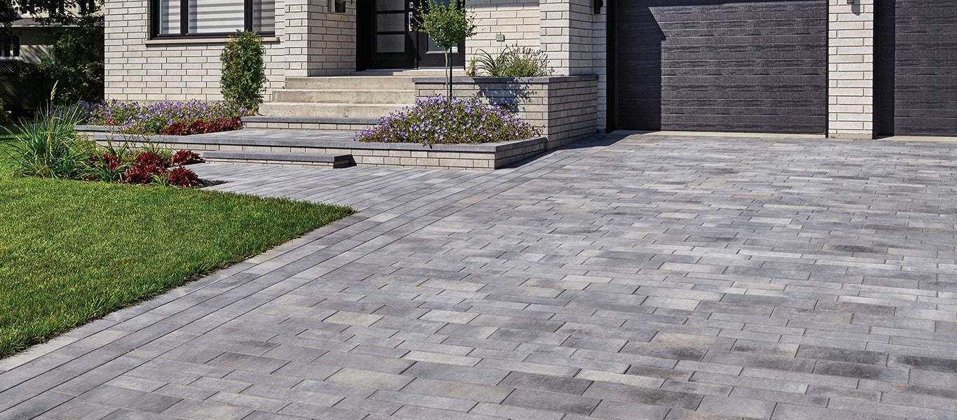A paved driveway made of grey stone tiles leading to the entrance of a modern home with a lawn on the side.