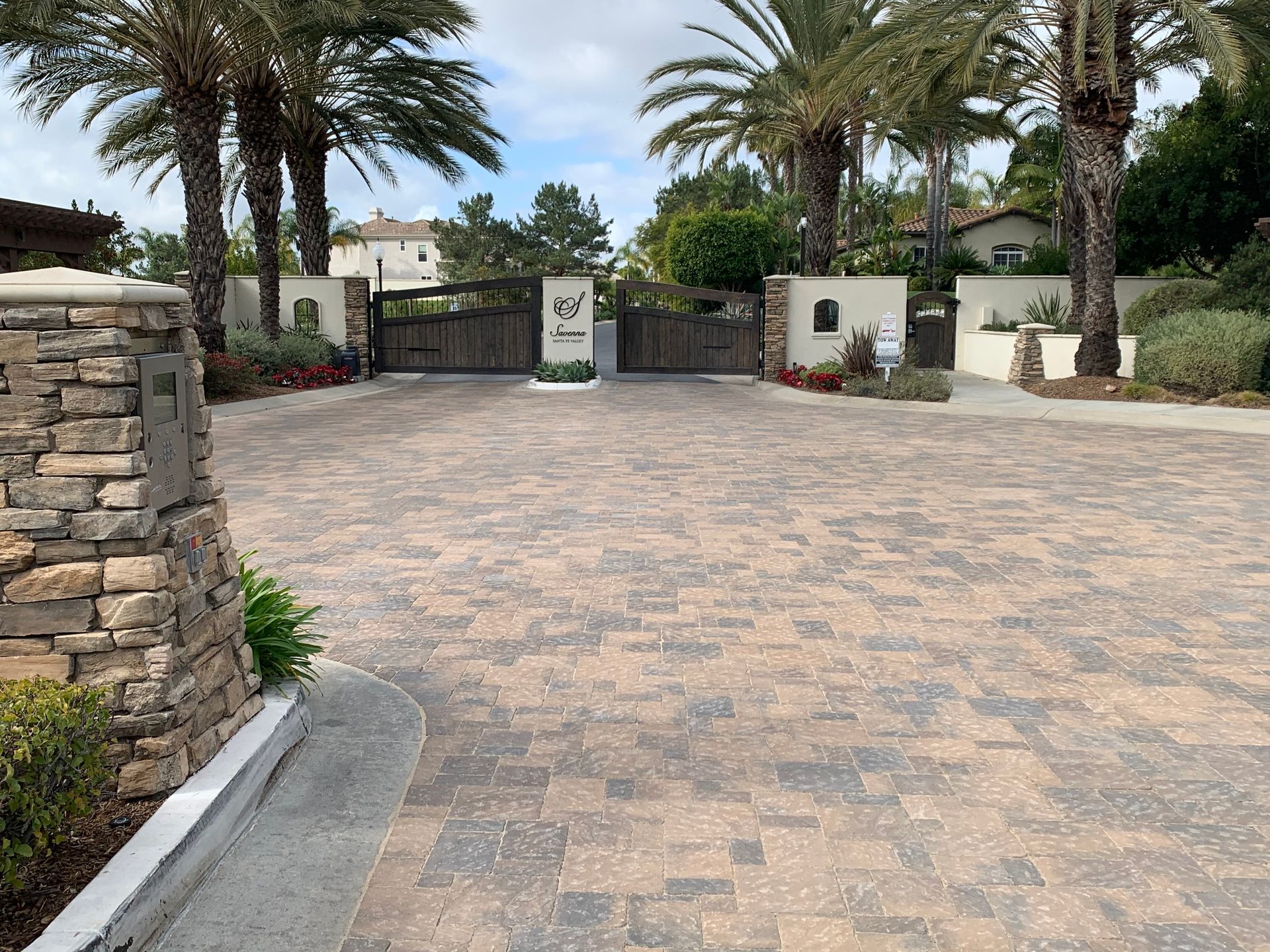 A stone-paved driveway leads to a gated entrance with palm trees.