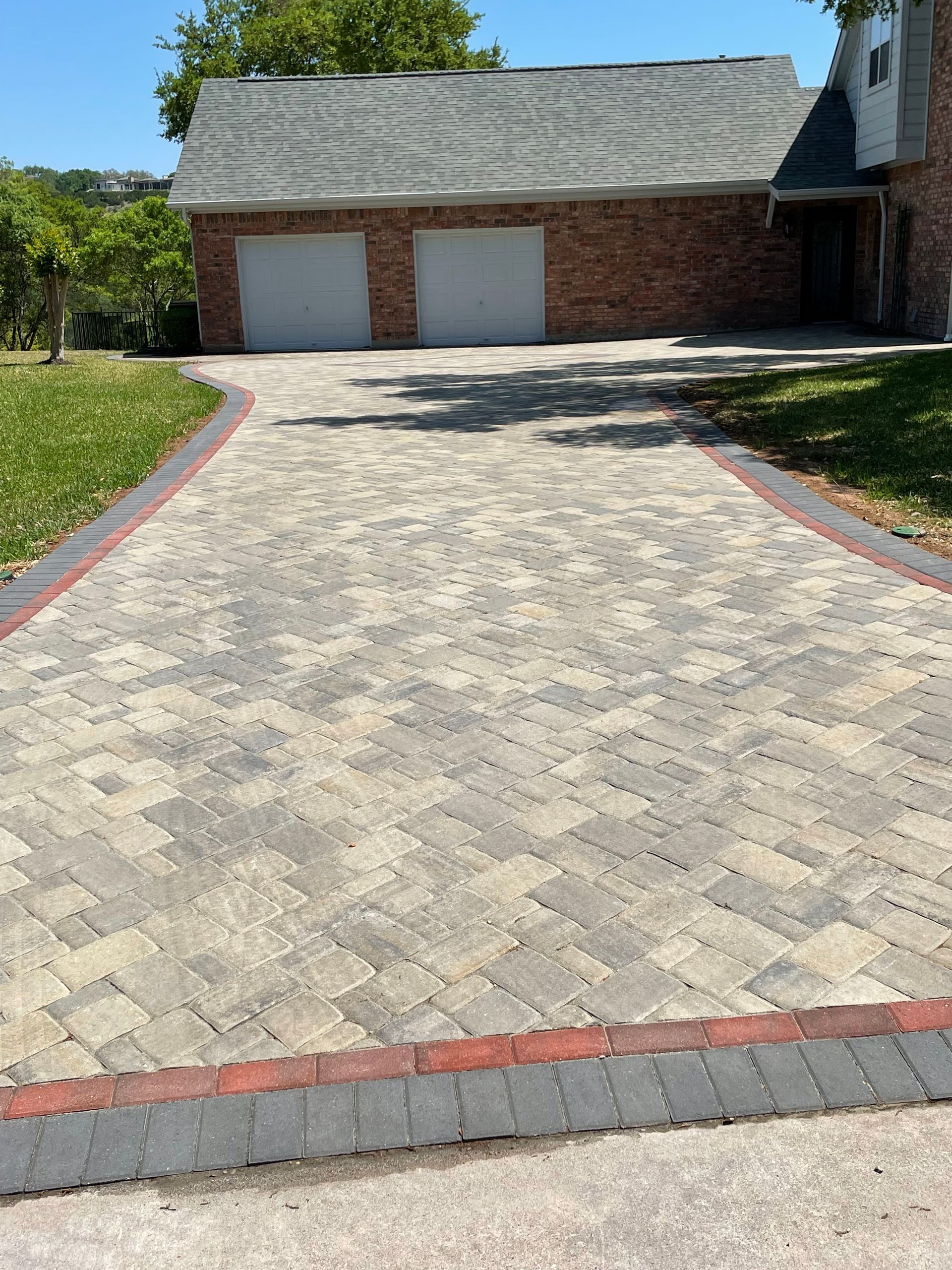 A paved driveway made of multi-toned grey and beige bricks with a red and charcoal border leading to a two-car garage.