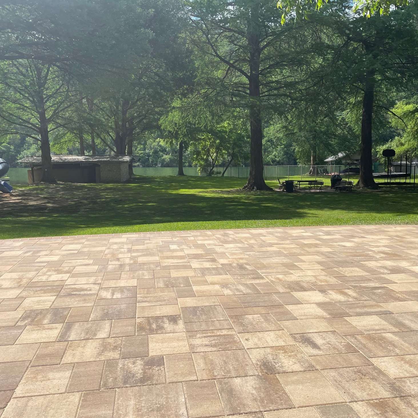 A tan paver patio overlooking a lush green grassy area with tall trees leading to a body of water.