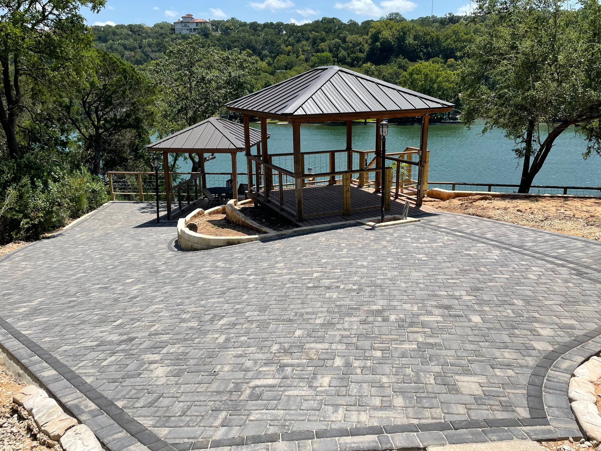 Two gazebos with dark metal roofs on a patterned stone patio overlooking a lake surrounded by trees.
