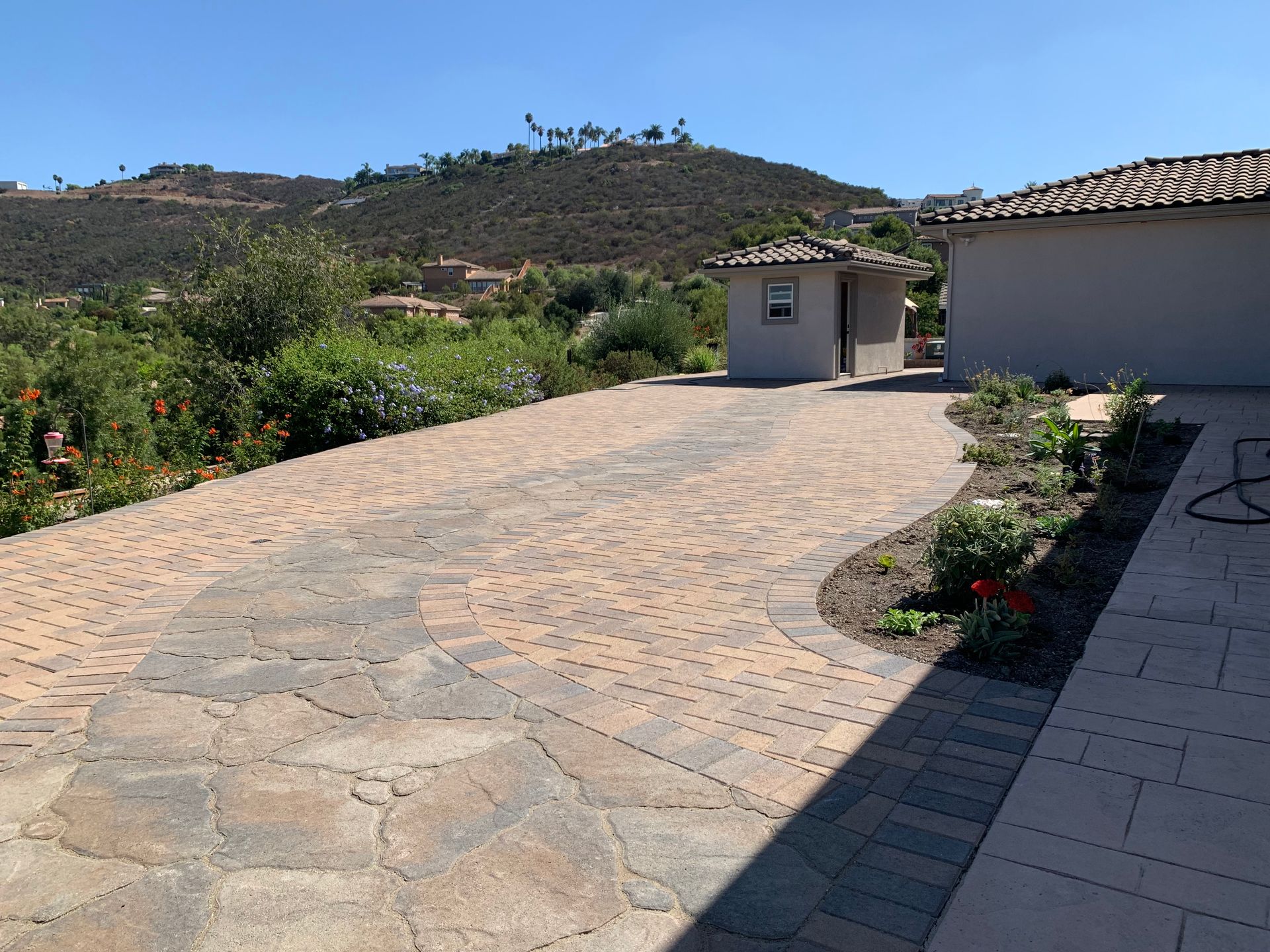 A curved, multi-toned stone paver driveway leads to a small detached structure in a sunny.