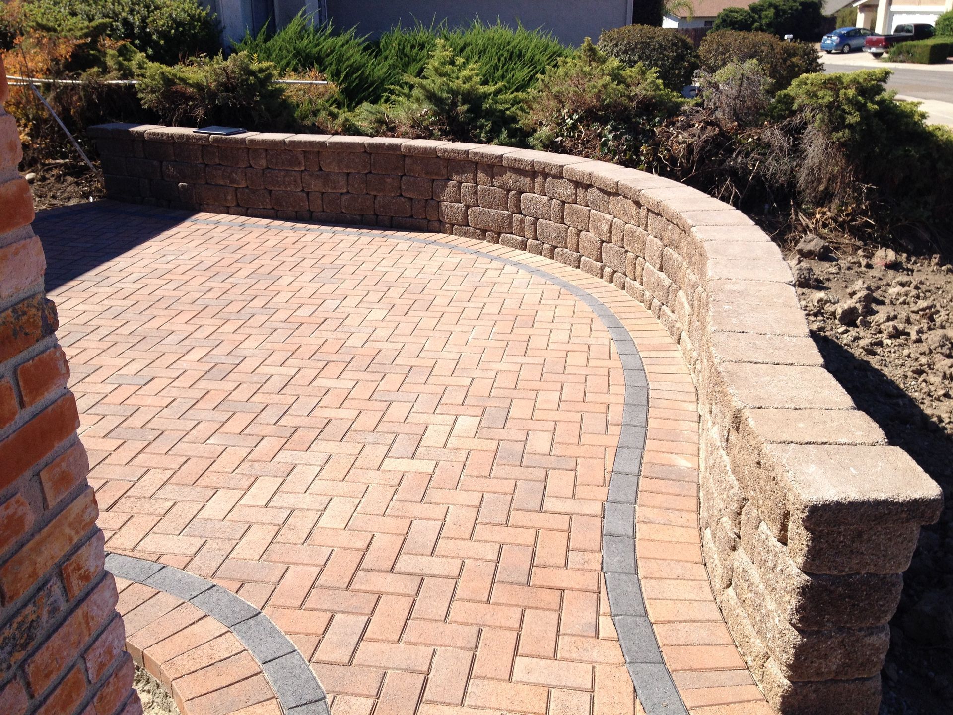 Curved retaining wall made of stacked stone blocks next to a herringbone-patterned paver patio in a sunny garden setting.