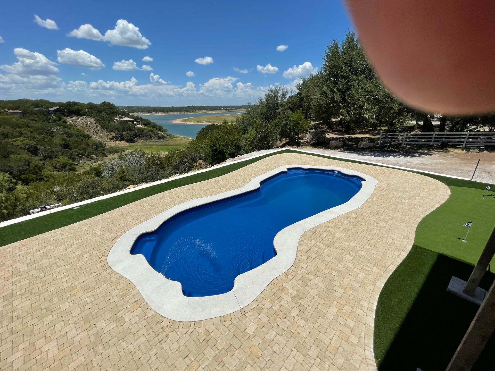 A blue, freeform swimming pool surrounded by a tan paver deck and artificial turf.