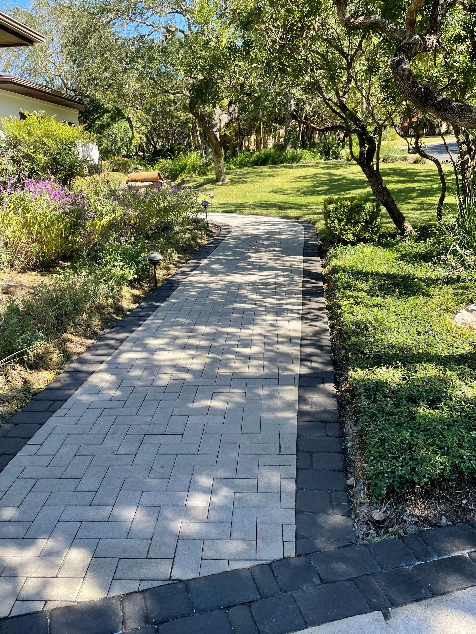 A straight stone walkway with a light-colored herringbone center and dark border, surrounded by grass and trees.