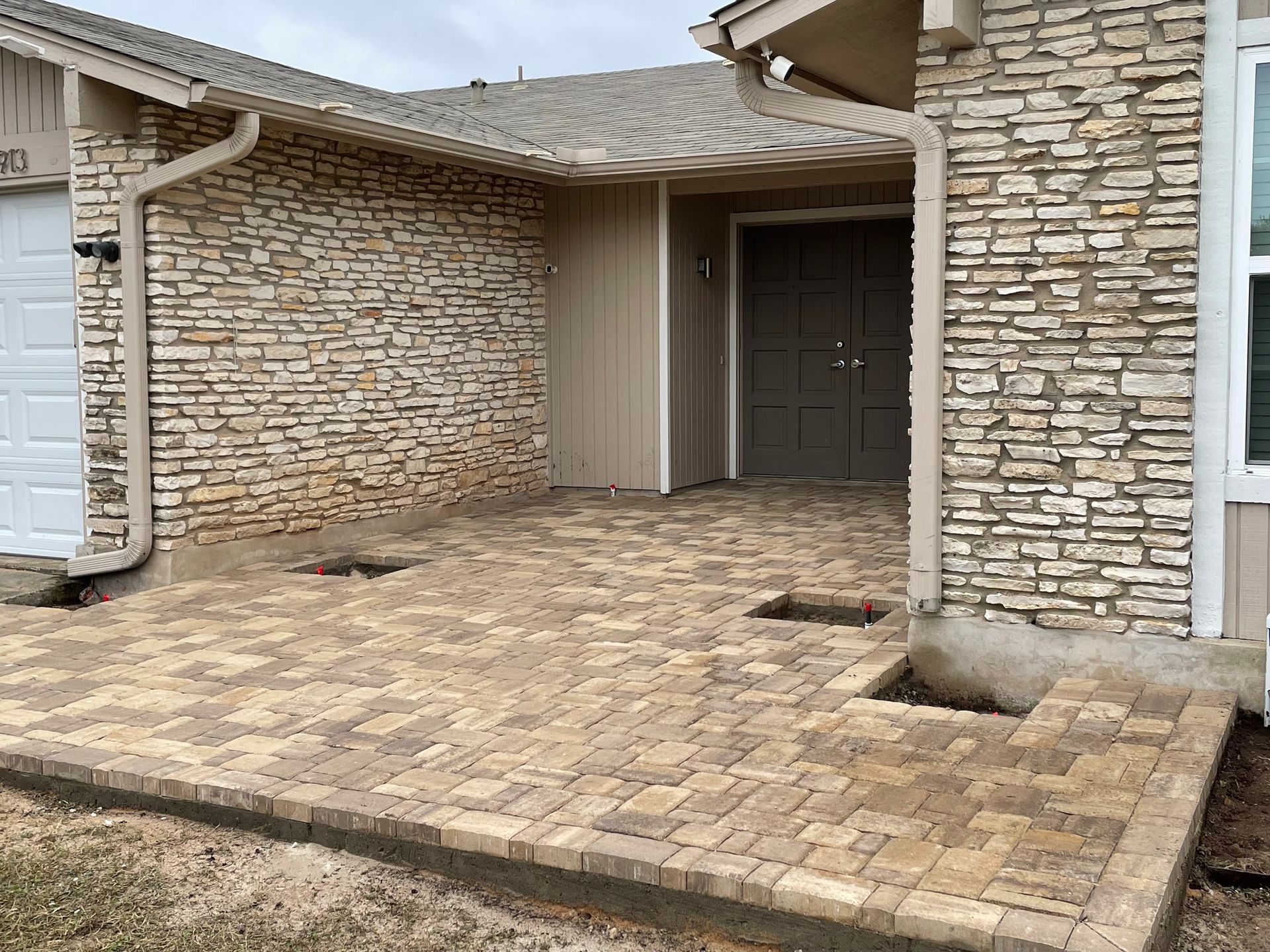 A newly installed beige paver patio in front of a house with tan stone siding and a dark double door.