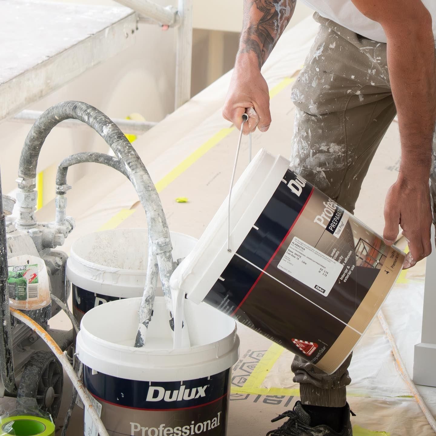 A Person Pouring White Paint From A Dulux Professional Bucket Into Another White Bucket In A Constru