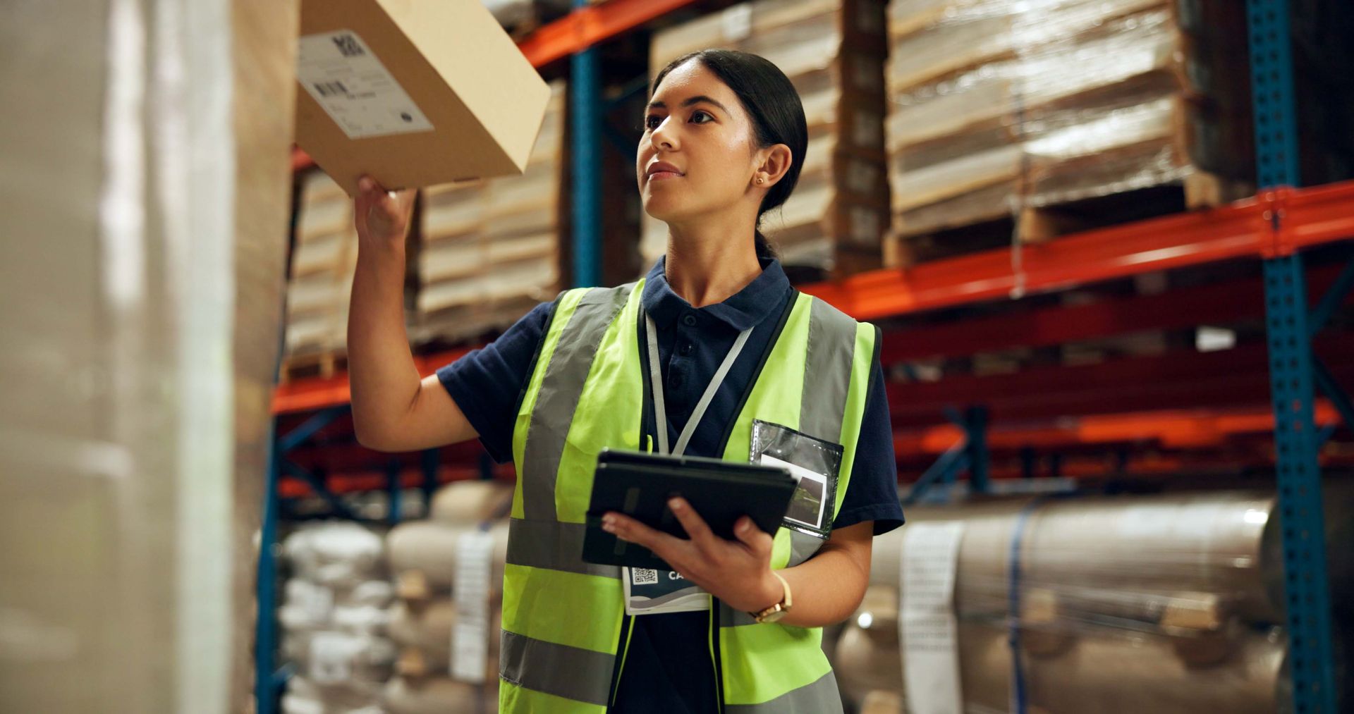 Warehouse worker in safety vest holding a package and using a tablet.