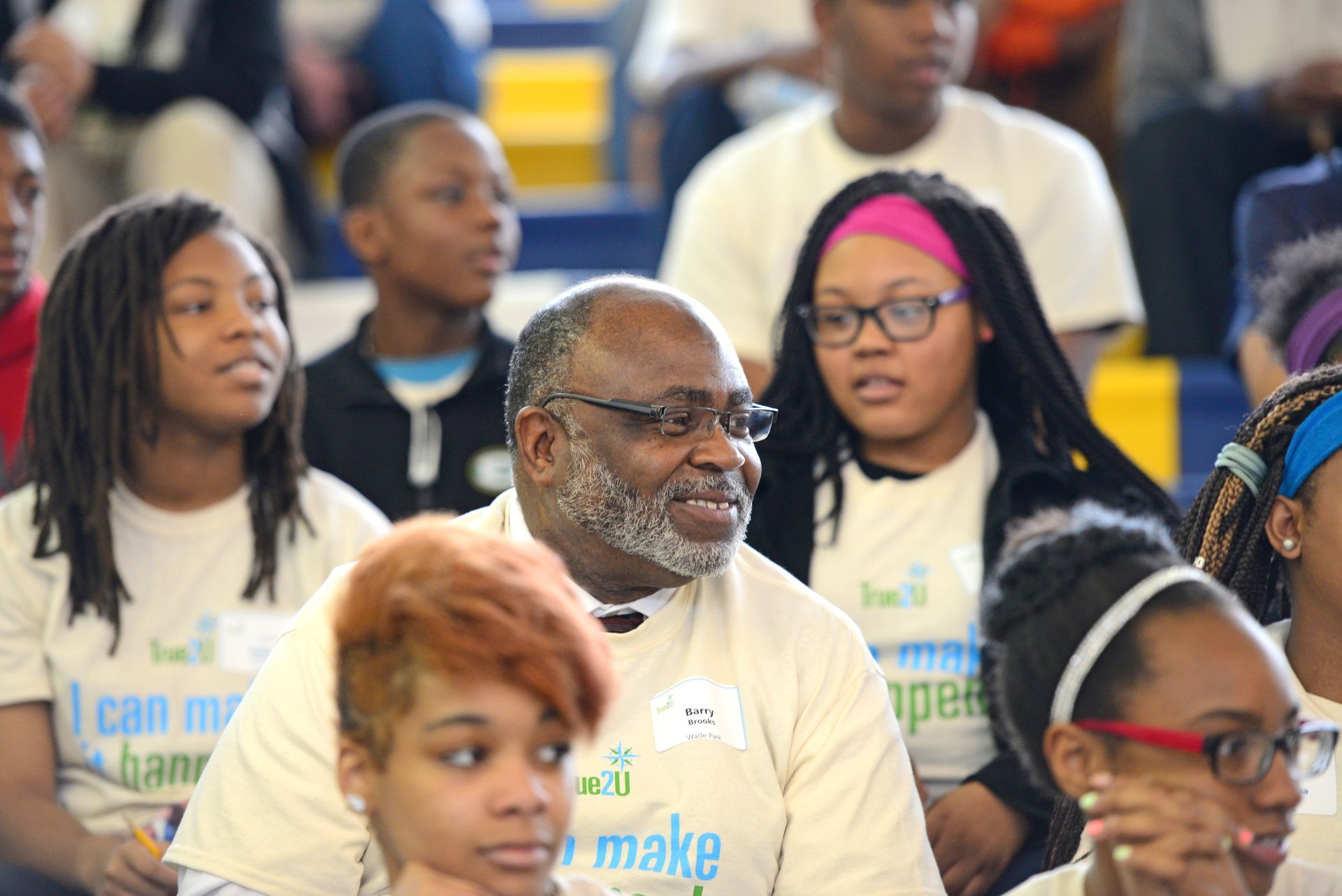 A man wearing a shirt that says i can make is sitting in a crowd of people