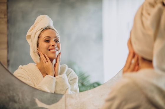 A woman with a towel wrapped around her head is applying a cream to her face.
