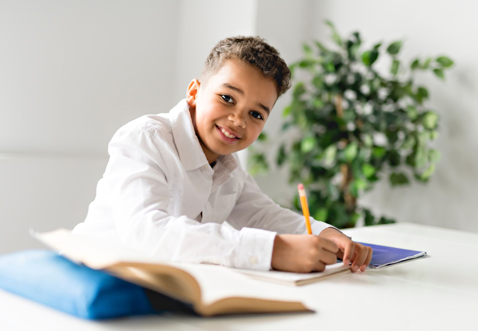 Boy writing in notebook, smiling, at a white desk with a book and plant in background.