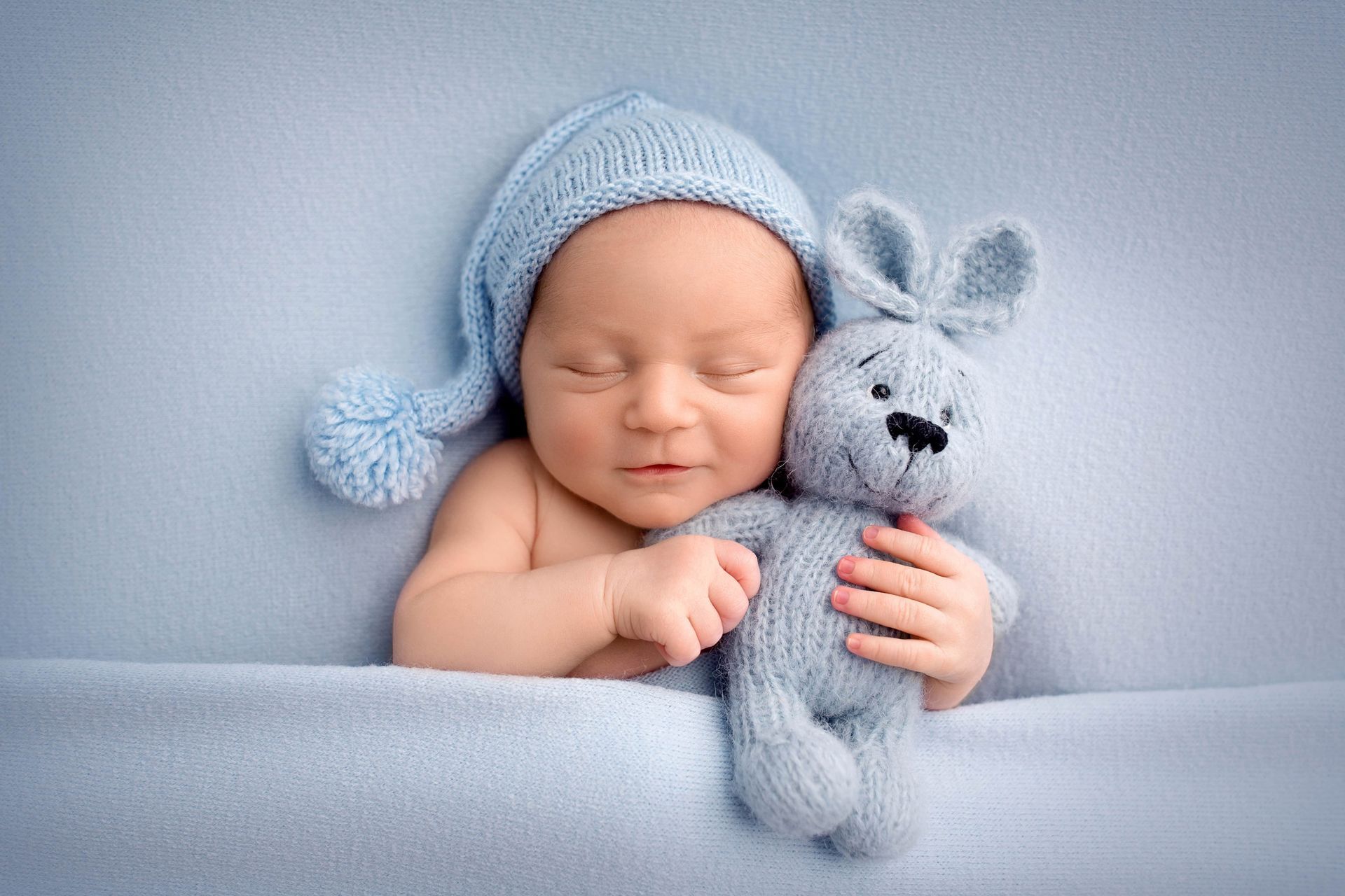 Baby wearing a blue knitted hat, cuddling a blue stuffed bunny, on a blue blanket.