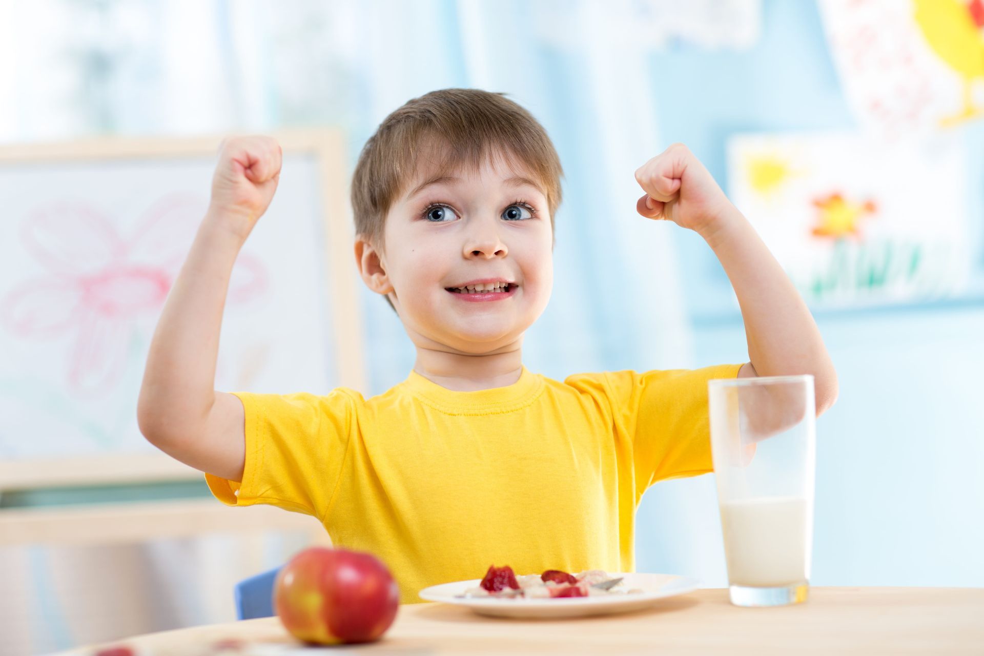 Boy with arms raised, smiling, at table with apple, strawberries, and milk.