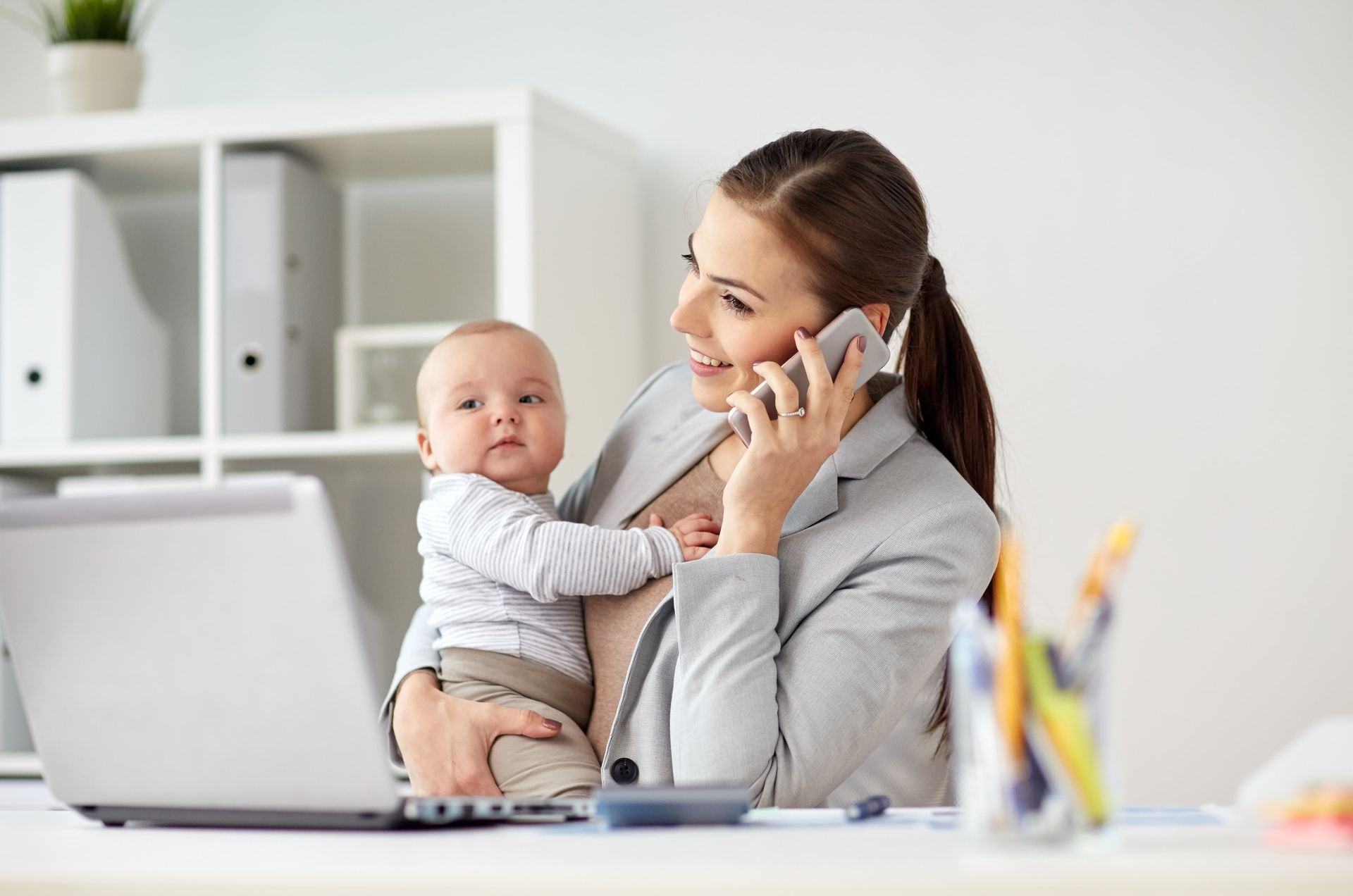 Woman holding baby, talking on phone, working at a desk with laptop and office supplies.