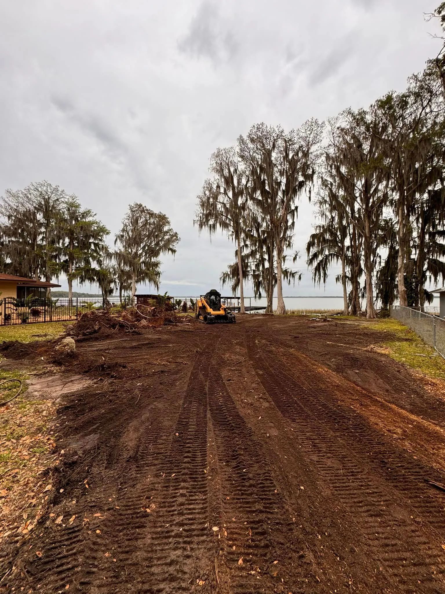 A construction site with a skid steer on muddy ground near trees and water, under a cloudy sky.