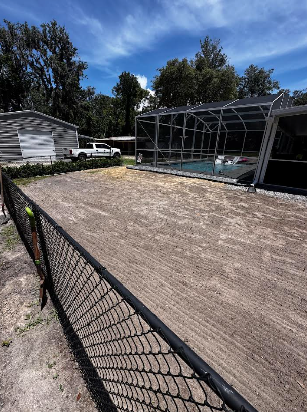 Black chain link fence frames a dirt yard, with a pool enclosure and small shed under a blue sky.