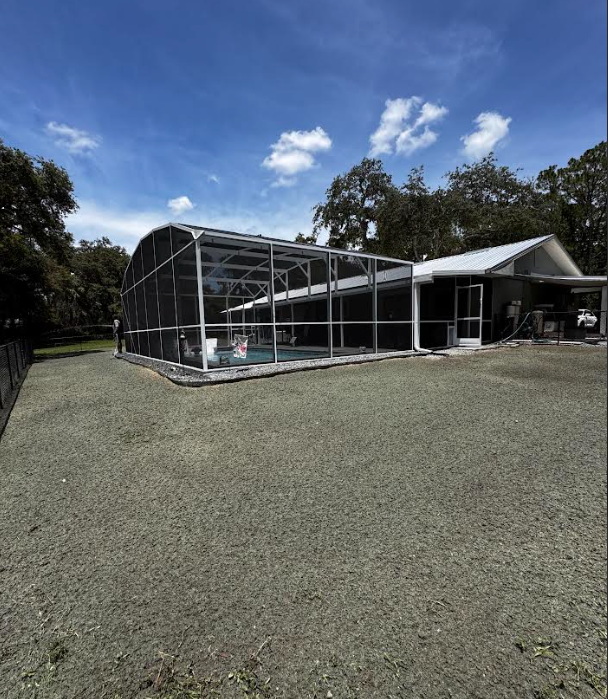 A backyard with a screened-in pool, a house with a white roof, and green grass under a blue sky.