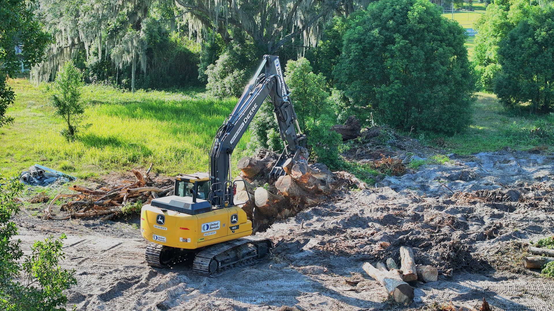 Yellow excavator removing cut tree logs in a grassy area.