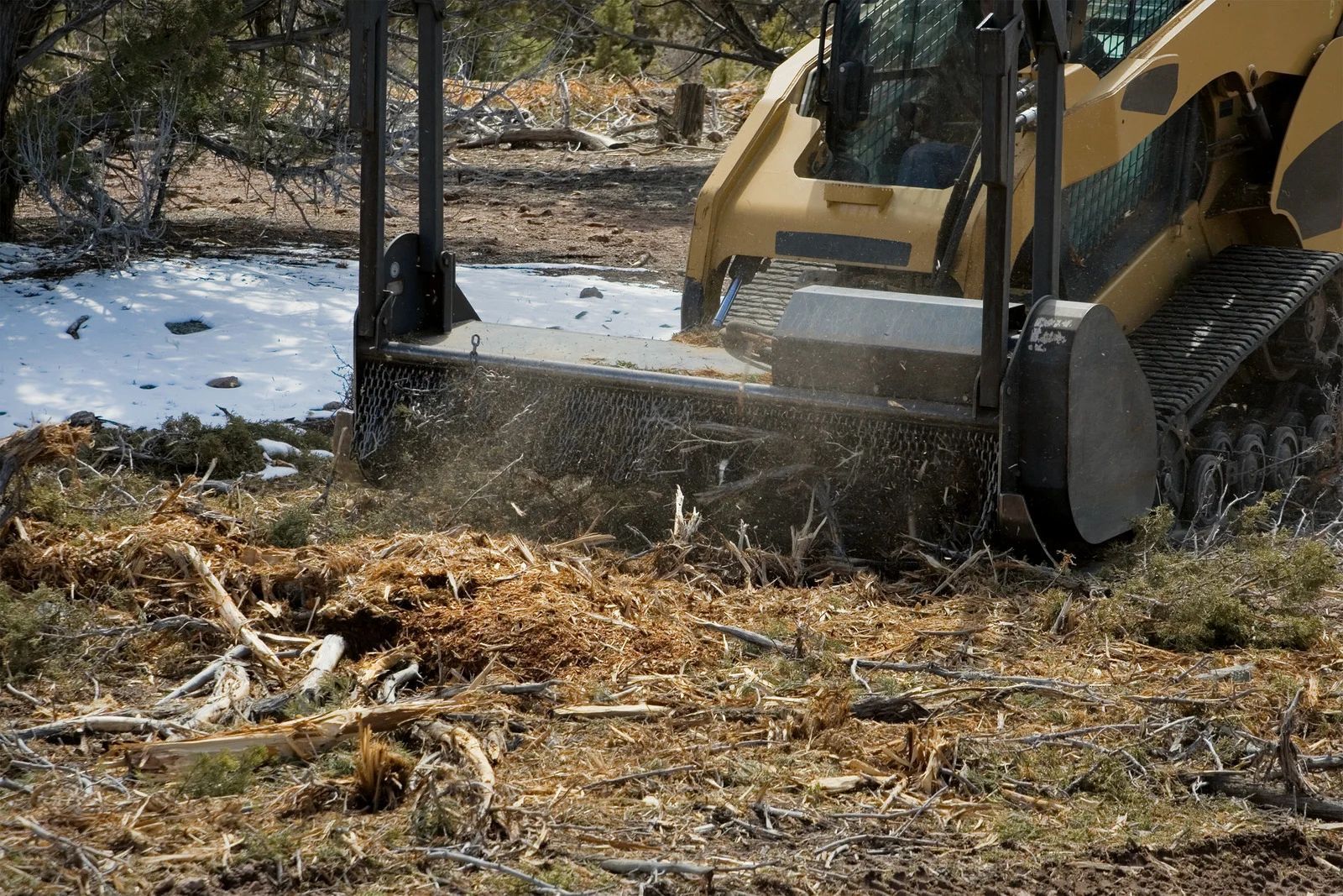 Yellow skid steer mulching brush and woody debris in a field with patches of snow.