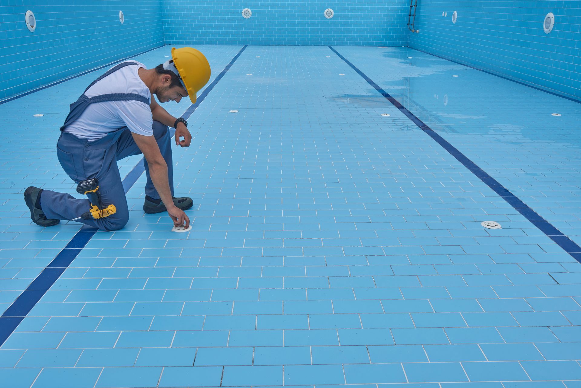 Person in overalls and hard hat inspecting a tiled swimming pool.
