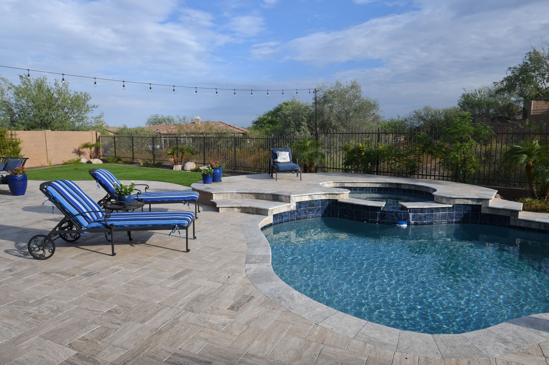 Backyard pool with blue lounge chairs, stone patio, and trees under a clear sky