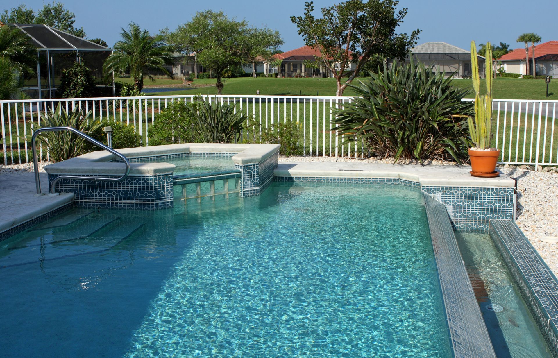 Pool with a built-in spa. Blue water, white fence, plants in pots and garden, sunny day.