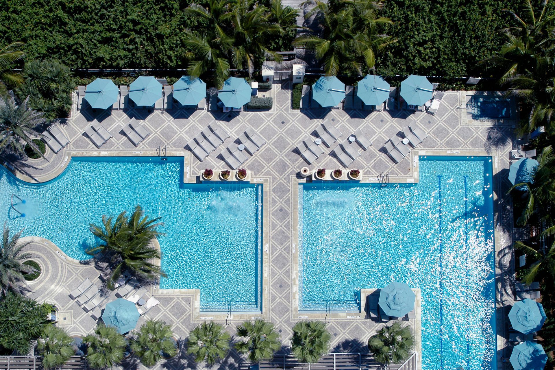 Overhead view of two blue swimming pools with lounge chairs, umbrellas, and surrounding greenery.