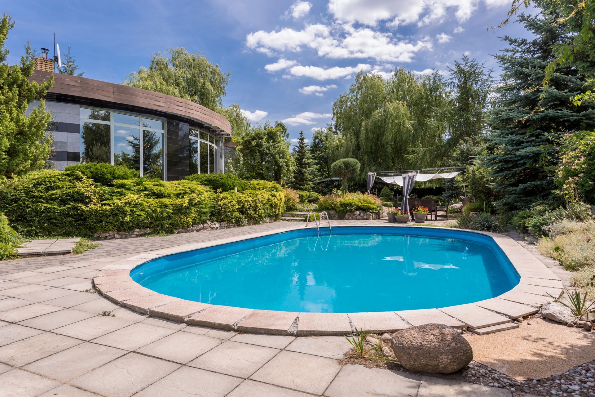 Swimming pool in a backyard with a house, greenery, and a partly cloudy sky.