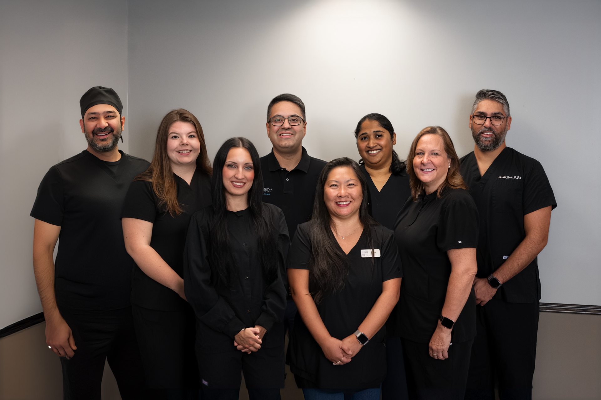 Group of eight people in black scrubs, smiling, posing in an office setting with a gray wall.